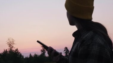 Günbatımında dağlarda akıllı telefonlardan beslenen aktif genç kadın parşömenlerinin silueti. Cep telefonu uygulama pazarı, podcast dinleme. Macera, doğa, rahatlama konsepti