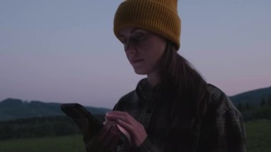 Genç, güzel bir bayan cep telefonu kullanıyor, sohbet ediyor, arkadaşlarıyla mesajlaşıyor, karanlıkta akıllı telefondan sosyal medyada haber yayıyor. Bereli yürüyüşçü kız. Arka plandaki karanlık gökyüzüne bak.