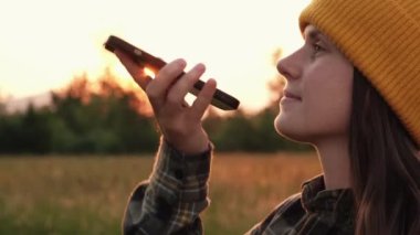 Bereli Hipster 'lı genç bir kadının arka plandaki destansı günbatımında cep telefonuyla ses mesajı gönderdiğini, genç bir öğrencinin otların üzerinde sesli mesaj araması yaptığını, ses arama kavramını kullandığını.