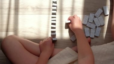 Close-up top view of little caucasian girl child builds lines with small dominoes sitting alone on warm floor at home. Concept domino effect in business, education, development fine motor skills