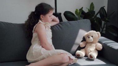 Portrait of upset lonely bullied little girl child looking away feels abandoned abused, sad unhappy small kid orphan sitting alone on grey sofa at home near teddy bear toy, charity adoption concept