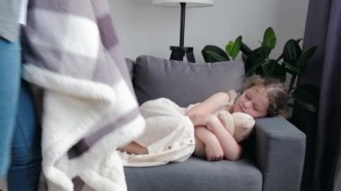 Caring mom covering sleepy little girl with blanket or plaid. Loving mother touching, caressing, comforting sleeping daughter. Child care concept. Cute peaceful kid taking nap on couch in living room