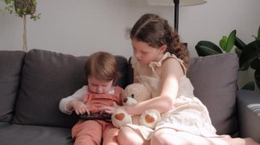Two cute relaxed little sisters sitting on cozy couch at home using mobile phone.Close up of pretty small daughters looking at device screen. Technology new generation addicted with gadgets concept