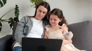 Caring young mum and small kid daughter watching video cartoons or playing game on laptop, mother teach little child girl online shopping make webcam video call looking at computer screen sit couch