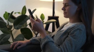 Young woman sit relax on sofa under night lamp in living room browsing surfing wireless internet on smartphone, girl rest on couch at home message text on modern cellphone, shopping online via website
