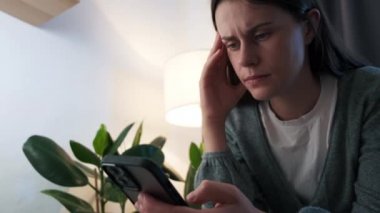 Worried young woman reading bad news in message on smartphone sit alone on sofa under light of lamp at home. Anxious girl feeling jealous upset holding mobile phone thinking of problem receiving sms