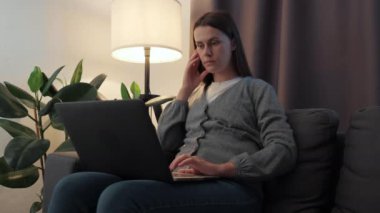 Pretty young woman typing on computer browsing internet working studying communicating online surfing web looking at screen. Smiling female using laptop notebook sit on couch under night lamp at home
