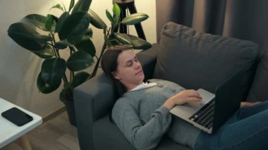 Relaxed young female typing on computer browsing internet working studying communicating online surfing web looking at screen. Cute girl using laptop notebook lying on sofa under night lamp at home
