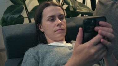 Close up of young woman lying on comfy couch under night lamp in living room browsing surfing wireless internet on smartphone, girl rest at home message text on modern cellphone. Shopping online