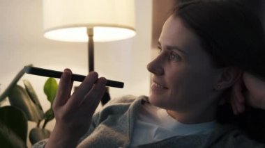 Close up of happy female sit on couch at night activating virtual assistant on mobile phone. Smiling young woman recording answering by audio message in social network for friend or using translator