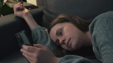 Lazy indifferent woman resting in couch under night lamp, using smartphone. Mobile phone user relaxing on sofa, browsing internet, using online, checking social media, suffering from digital addiction