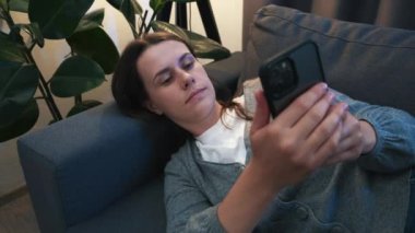 Smiling young woman lying on cozy sofa under light of lamp in living room browsing surfing wireless internet on smartphone, girl rest on couch at home message text on modern cellphone. Shopping online
