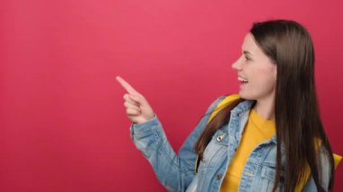 Happy young brunette woman with yellow backpack pointing on copy space for promotion content, posing isolated over red color background in studio. Education in high school university college concept