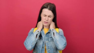 Unhappy young student girl has neck pain, tilts head, tired after having classes at university, returns home fatigue, wearing denim jacket and rucksack, isolated over red color background in studio