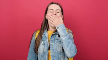 Portrait of lazy young student girl with denim jacket and yellow backpack making sleep gesture in adorable expression, isolated over red background. Education in high school university college concept