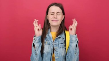 Cute young girl teen student in denim jacket and yellow backpack keep finger cross making with eyes closed, posing isolated on red background wall. Education in high school university college concept