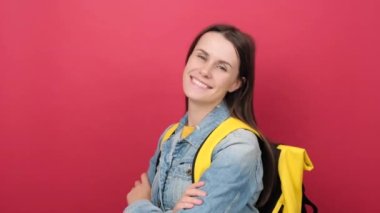 Portrait of smiling happy fun teen student girl wearing denim jacket and yellow backpack, look camera isolated on pastel red color background wall in studio. Education in university college concept