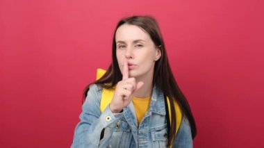 Portrait of young student woman doing silence gesture, wearing denim jacket and yellow backpack, posing isolated on red color background in studio. Education in high school university college concept