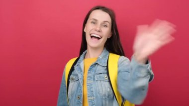 Friendly young student woman saluting with hand, happy expression, wearing denim jacket and yellow backpack, posing isolated over red color background in studio. Education, technology, online concept