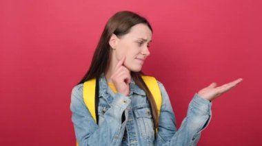 Pensive young student woman having doubts, wearing denim jacket and yellow backpack, posing isolated over red color background wall in studio. Education in high school university college concept