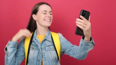 Smiling young student woman in denim jacket and yellow backpack doing video call in mobile phone, posing isolated over red color background wall in studio. Education school university college concept