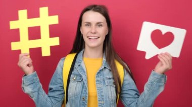 Pretty girl teen student with backpack holding yellow hashtag and heart icon, wears backpack and denim jacket, isolated over red color background in studio. Education, technology, online concept
