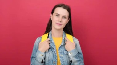 Happy young student woman wearing yellow backpack and denim jacket beckoning come here gesture with hand inviting welcoming happy and smiling, posing isolated over red color background wall in studio