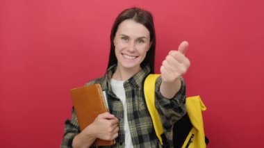 Portrait of happy beautiful young student woman with yellow backpack holding notebooks presenting and inviting to come with hand, posing isolated on red studio background wall. Education concept