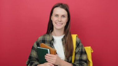 Portrait of young smiling happy girl teen student wear shirt and yellow backpack holding books, posing isolated over red color background in studio. Education in high school university college concept