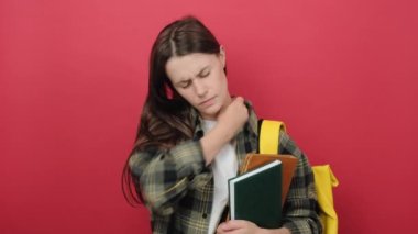 Upset young student woman suffering from pain in shoulder for having made an effort, wears yellow backpack, posing isolated on red studio background wall. Education in university college concept