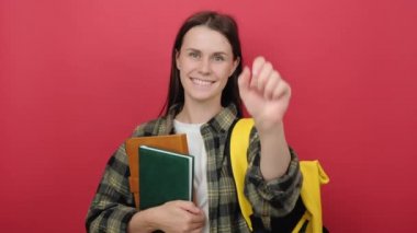 Portrait of happy young student woman with yellow backpack holding books and counting with fingers, posing isolated over red color background wall in studio. Education in university college concept