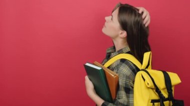 Pensive young student woman with yellow backpack and two books on back position looking back while scratching head, isolated on red color background in studio. Education in university college concept