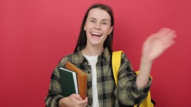 Portrait young joyful woman in shirt with backpack, looking at camera, waving her hand for greeting, meeting with friend, isolated on red studio background. Education at University College High School