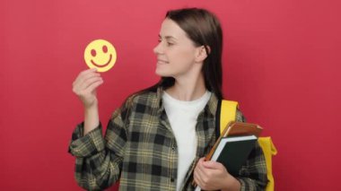 Portrait of young woman student holding small yellow paper happy emoticon, wears backpack, posing isolated over red color background in studio with copy space for promotion content. Education concept