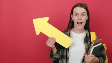 Smiling young student woman wears a shirt and backpack holding yellow arrow pointing on copy space for promotional content, isolated on red background wall. Education in university college concept