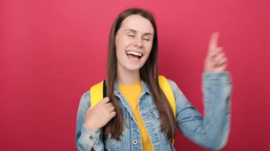 Cheerful smiling young brunette woman with yellow student backpack intending to realizes solution while lifting finger up, posing isolated over red color background wall in studio. Education concept