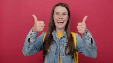 Happy smiling girl teen student wears denim jacket and yellow backpack showing thumbs up like gesture, posing isolated over pastel red studio background wall. Education in university college concept