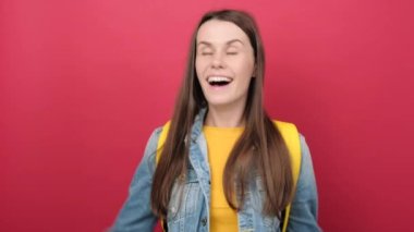 Cheerful young student girl in denim jacket and yellow backpack celebrating victory in winner position, posing isolated on red studio background. Education in high school university college concept