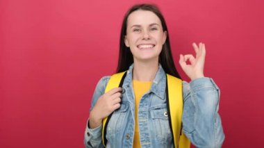 Happy beautiful young woman student with yellow backpack showing OK or zero gesture, wears denim jacket, posing isolated over red color background in studio. Education in university college concept