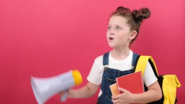 Smiling leader little kid school girl 6-7 years old with yellow backpack holding two books and scream in megaphone isolated on pastel red color background wall in studio. Childhood education concept