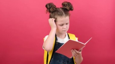 Pensive kid girl holding notebook gesture raises finger came up, good idea, positive looking at camera, wears t-shirt and yellow backpack, posing isolated over red color background wall in studio