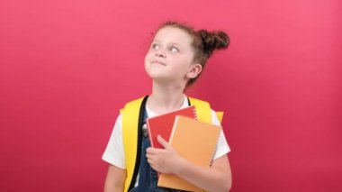 Portrait of dreamful little kid school girl 6-7 years old in t-shirt and backpack hold notebooks keep finger crossed, making wish eyes closed, isolated on red background. Childhood education concept