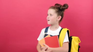 Portrait of school kid girl 6-7 years old in white t-shirt and yellow backpack holding two notebooks pointing fingers on workspace isolated over red background studio. Education lifestyle concept