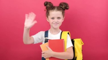 Portrait of happy smiling kid girl with yellow backpack and two notebooks ready to study at school waving hello, posing isolated over pastel red color background wall in studio. Education concept