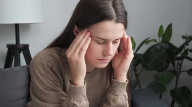 Close up of stressed young caucasian woman massaging temples, suffering from headache or thinking of difficult problem solution looking in distance, feeling doubtful unhappy alone sit on sofa at home