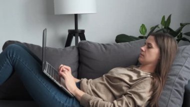 Relaxed young brunette woman lying on comfy sofa in living room, typing on laptop, creating text for personal blog in social network. Girl working on new business idea at free leisure time at home