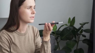 Close up of beautiful young woman listening pleasant news in audio message, recording response using cellphone application. Caucasian female communicating distantly by voicemail sit on sofa at home
