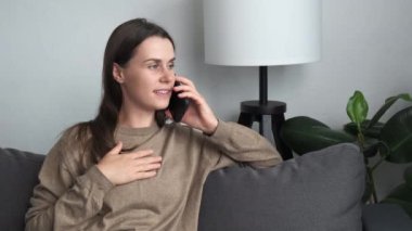 Pretty young caucasian woman sitting on cozy couch in cozy living room alone holding modern device having pleasant conversation on smartphone. Personal talk use modern wireless technology concept
