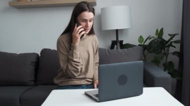 Cute young caucasian woman professional talking on phone using laptop sit on couch at home desk, focused female customer make mobile call confirming online website shopping order delivery concept