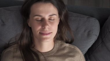 Close up of happy tranquil young brunette woman leaning on comfortable couch with closed eyes, sleeping breathing fresh humidified conditioned air. Cute beautiful girl napping relaxing alone at home